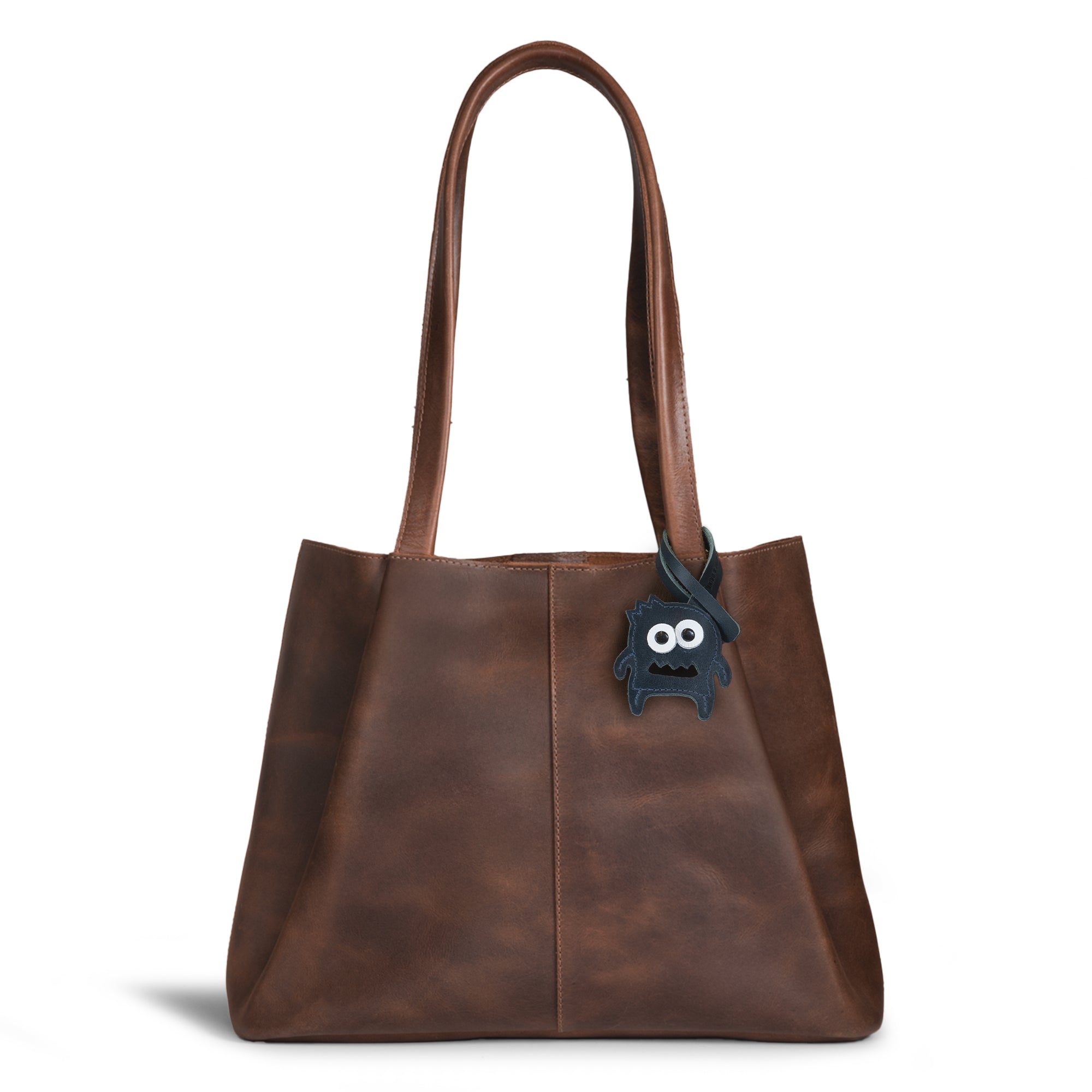 Hide & Drink Bag Charms attached to a trendy brown handbag, showcasing a skull design entwined with floral elements, perfect for personalization. This accessory serves as a purse charm for tote & backpacks crafted from durable full grain leather. Every piece reflects StockyardX's dedication to small batch production, with hand cut, hand sewn artistry and fire branding, handmade in La Antigua Guatemala & Oaxaca Mexico.