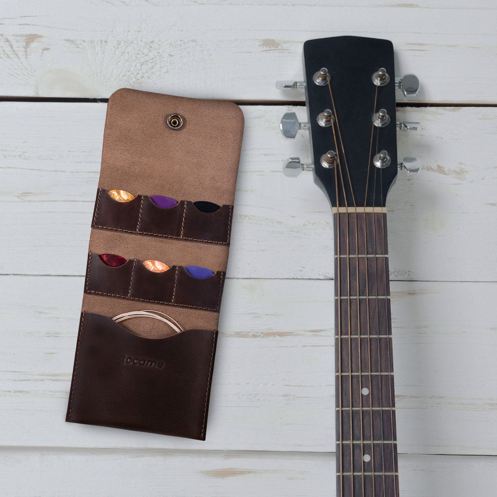 tocame's Guitar String Case with 6 Slots for Picks displayed next to a guitar, illustrating its design and how it complements musical gear. This Guitar String Case with 6 Slots for Picks serves as an ideal Guitar String Holder for Electric & Acoustic Guitar, crafted from Full Grain Leather and features artisan handmade quality from La Antigua Guatemala.