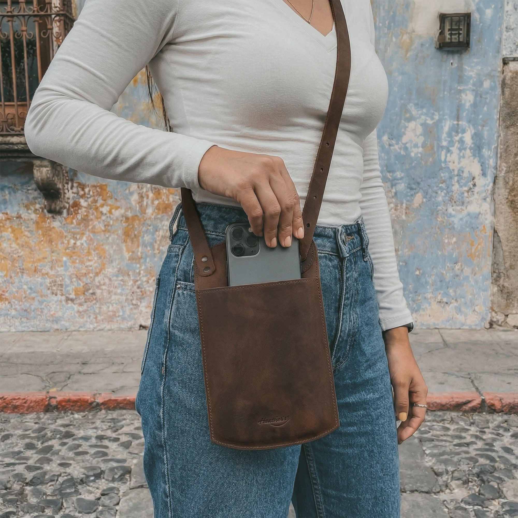 Lifestyle image of a person holding the Phone Purse with Adjustable Strap by Heather's, demonstrating its use as a convenient crossbody bag. This full grain leather bag integrates a horizontal cellphone design with an adjustable neck strap and card slot. Artisan crafted in La Antigua Guatemala, it reflects the brand's commitment to small batch production and handmade quality.