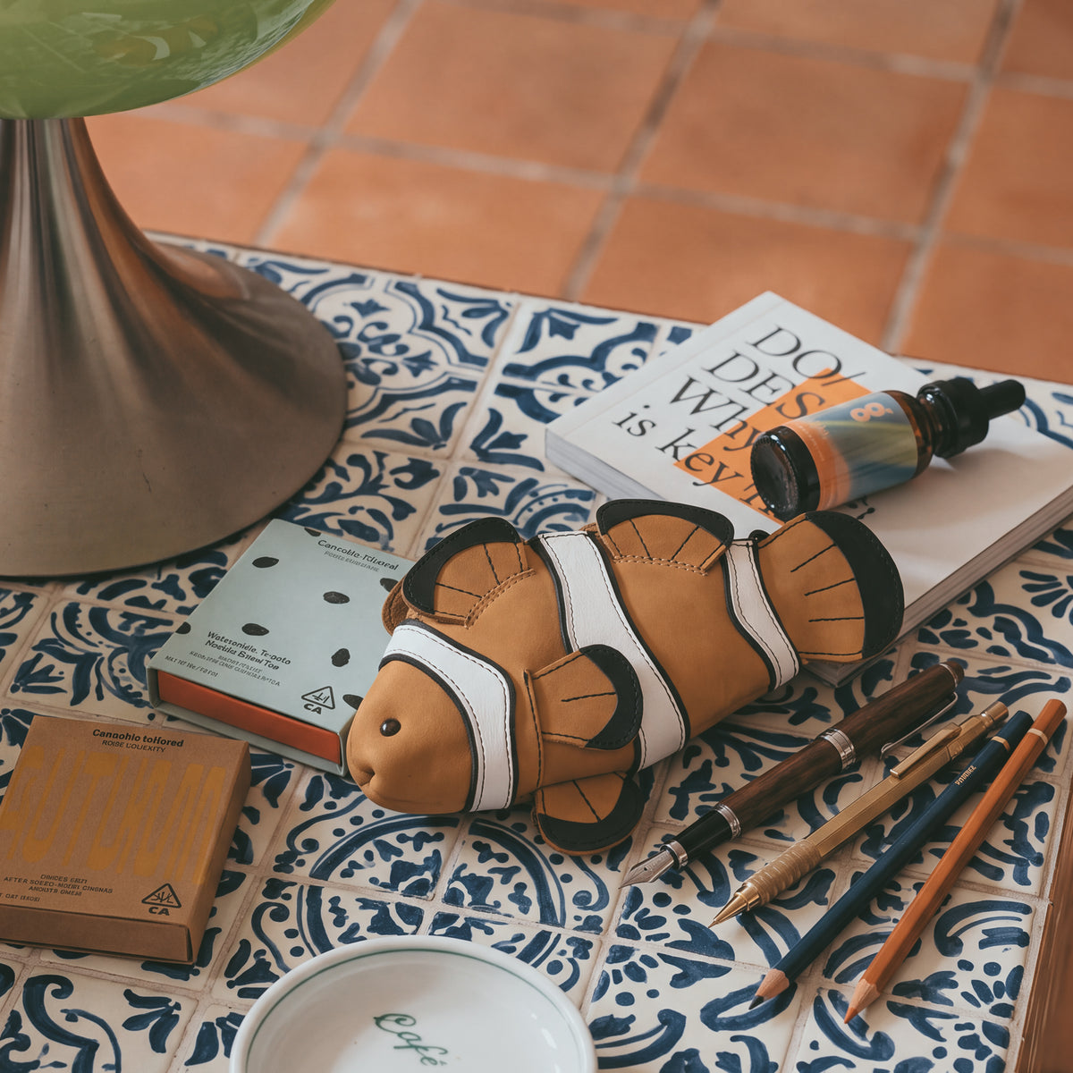 Tudi Frudi Leather Clownfish Pouch displayed on a patterned tabletop beside art supplies, books, and a drink, highlighting its use as a Leather Clownfish Pencil Pouch, Small Pen Organizer, Zippered Holder Wallet for Coins, Change Purse, Money Pouches for Men and Women, Saving Box, Cable Holder, Full Grain Leather, StockyardX, Small Batch Production, Hand Cut, Hand Sewn, Fire Branded, Artisan, Handmade in La Antigua Guatemala & Oaxaca Mexico.