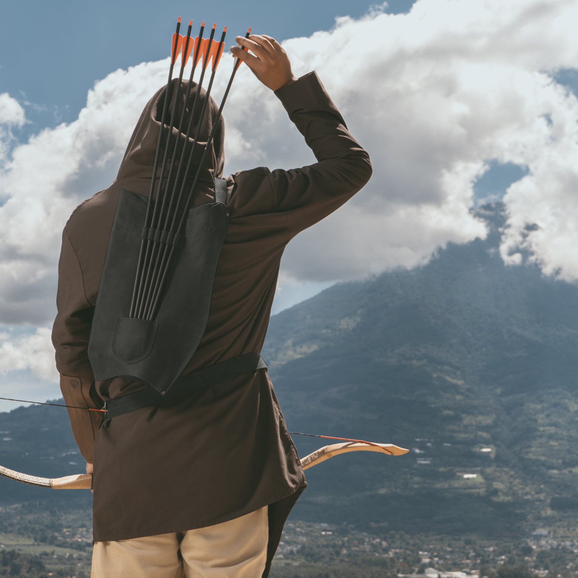 An archer donning the black Archery Bow Carrier with Arrow Slots by Valhalla Gear, set against a stunning mountain landscape. This full grain leather carrier with an adjustable strap is designed for shooting, training, and cosplay, highlighting artisan craftsmanship.