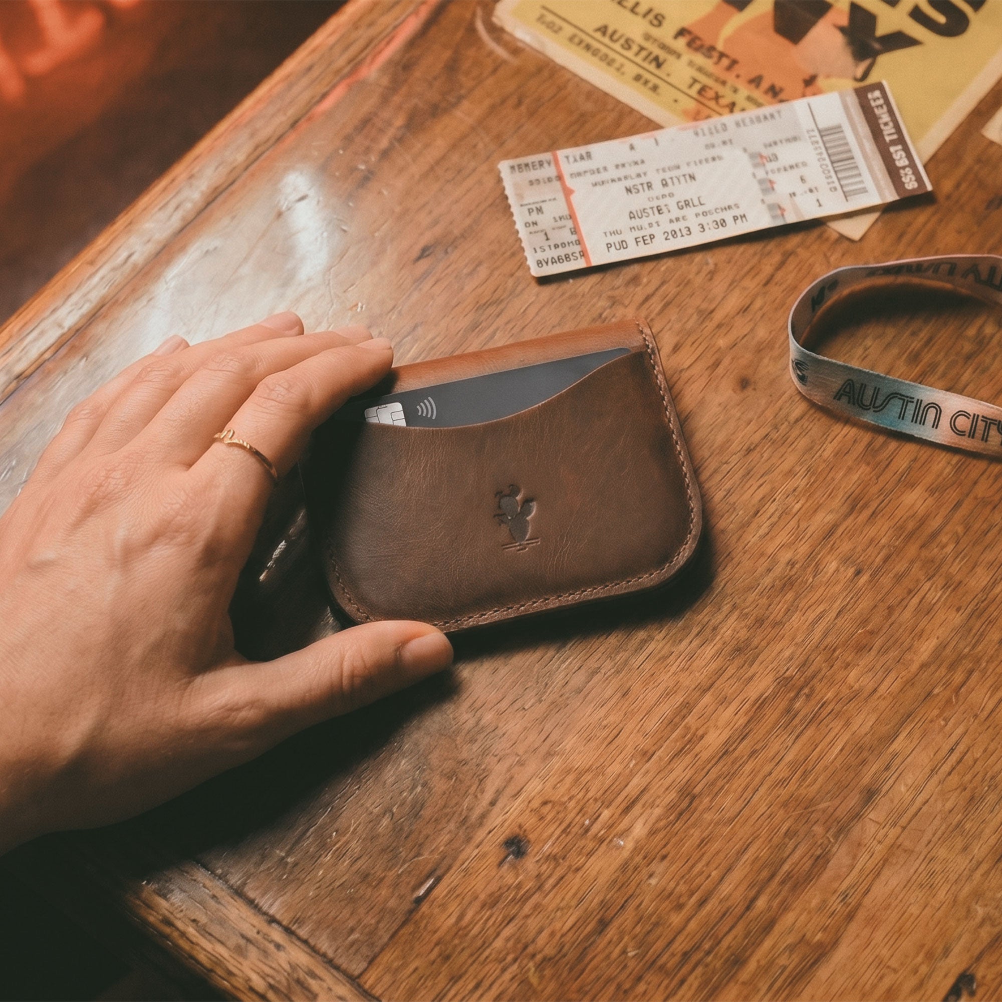 Austin Genuine The Magnolia Card Wallet resting on a wooden table beside travel tickets and accessories, with a card visible inside. The Magnolia Card Wallet is Full Grain Leather, made through StockyardX Small Batch Production, Hand Cut and Hand Sewn by an Artisan, and Handmade in La Antigua Guatemala & Oaxaca Mexico with Fire Branded detailing.