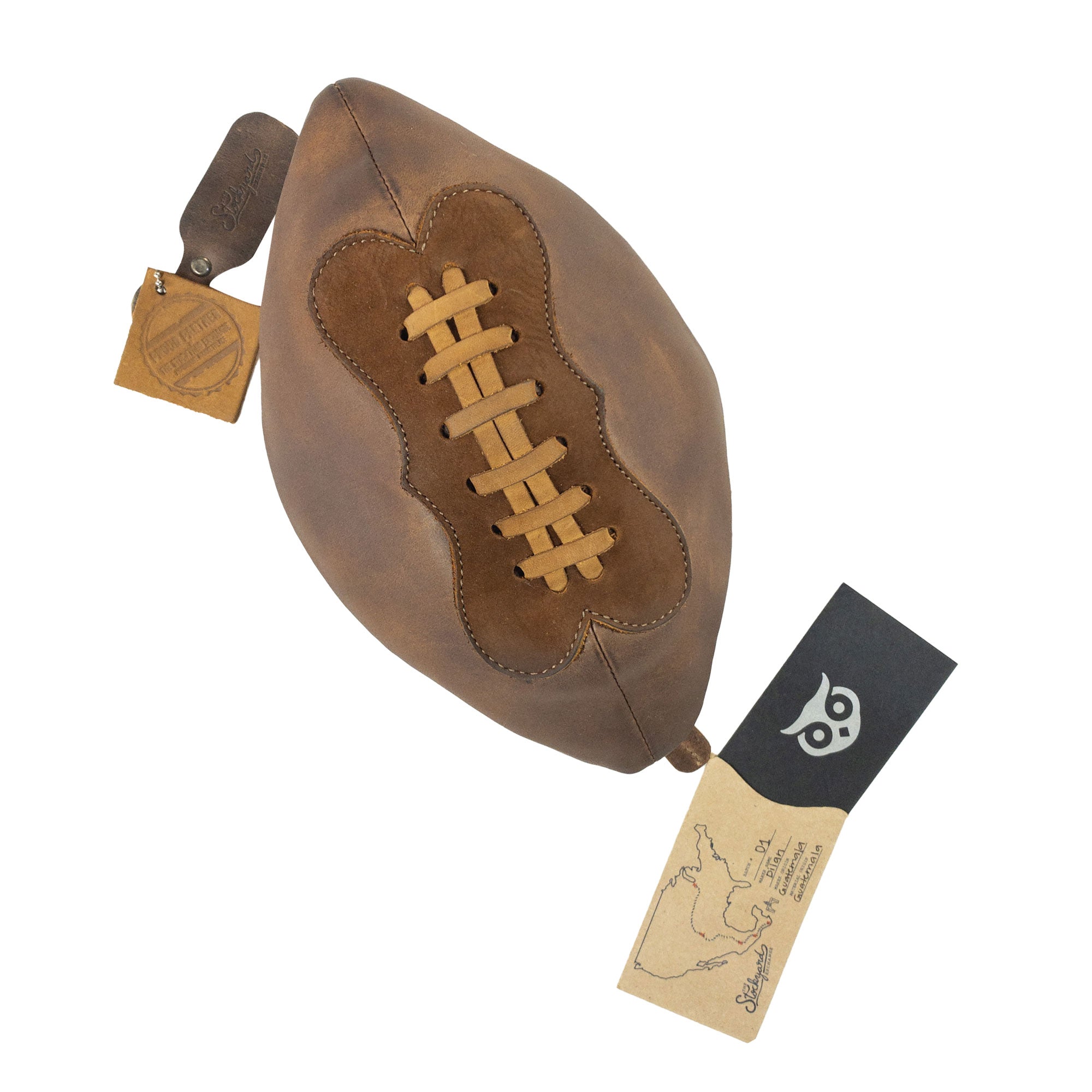 Football Toiletry Bag by Hide & Drink showcased with tags, highlighting its unique design and artisan quality. This Gameday Football Dopp Toiletry Bag is perfect for travel organization and grooming essentials, made from durable full grain leather. Featuring hand cut, hand sewn, and fire branded details, this handmade piece exemplifies craftsmanship from La Antigua Guatemala and Oaxaca Mexico.
