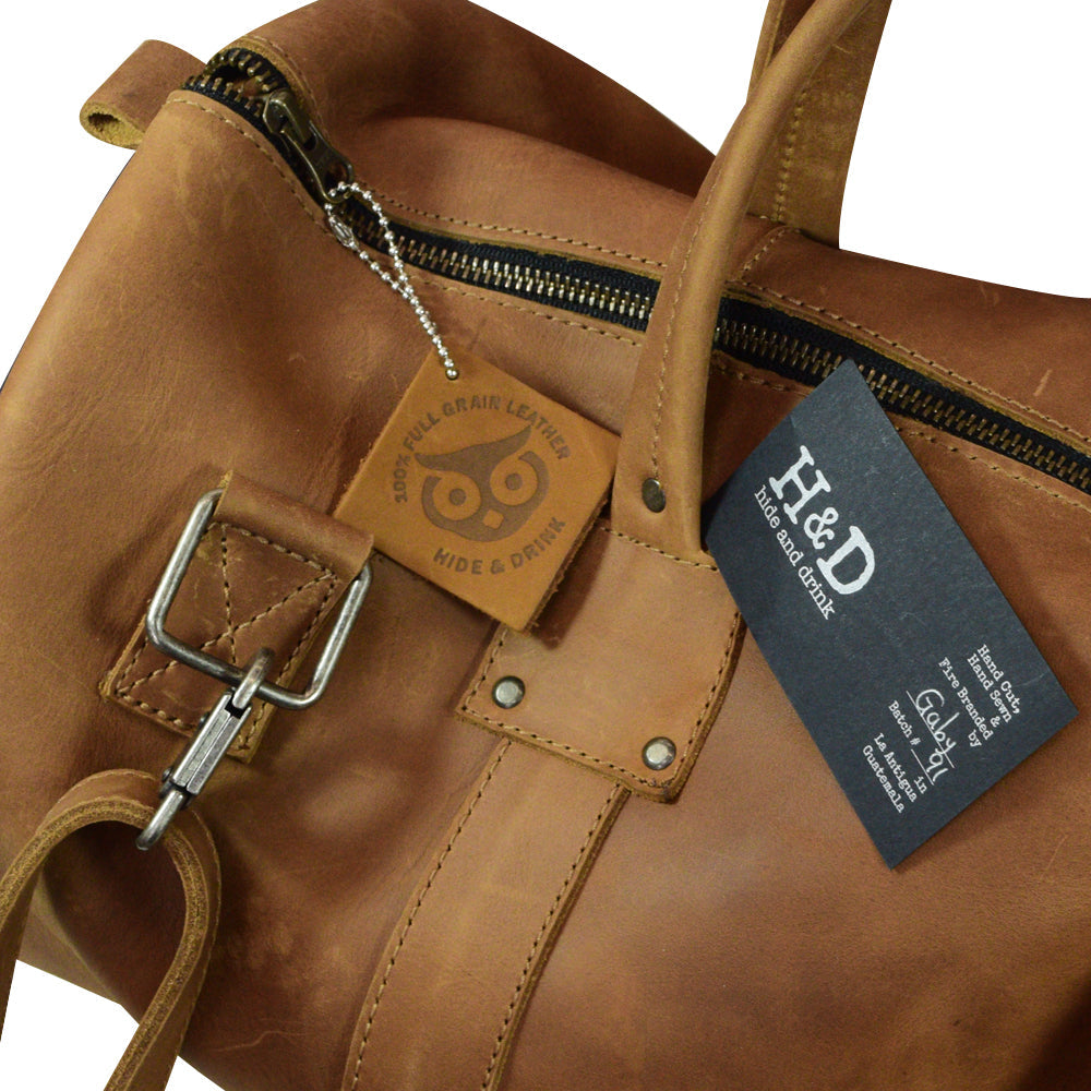 Overview of the Classic Duffle Bag by Hide & Drink in tan leather, showcasing spaciousness and practical design with zipper closure and adjustable strap. This Full Grain Leather Classic Duffle Bag serves as a stylish Messenger Bag ideal for travel, handmade in La Antigua Guatemala & Oaxaca Mexico.