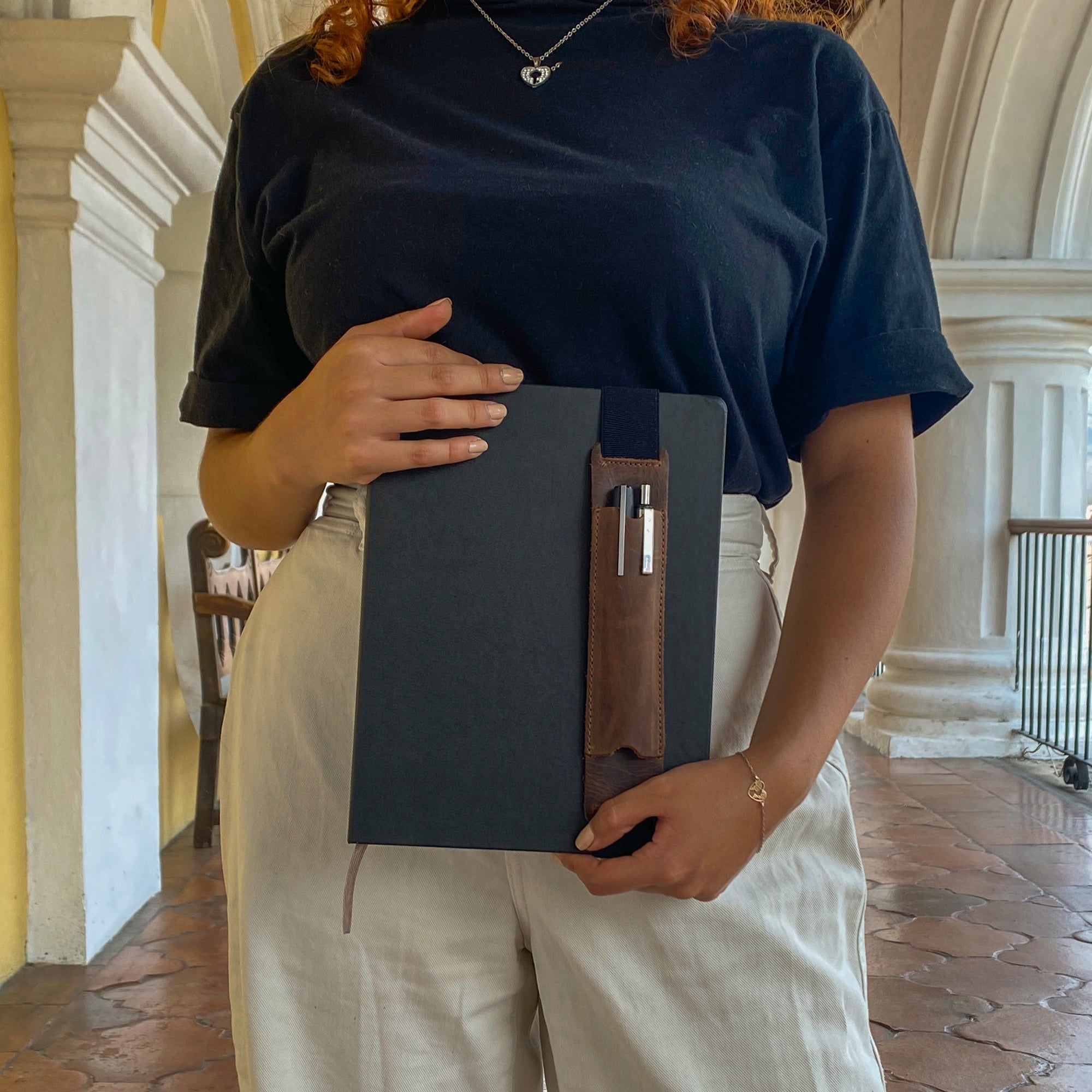 Person holding Hide & Drink Pen Holster, showcasing full grain leather and elastic band design. Perfect for 6 to 7 inch notebooks, this pen case is a stylish accessory, crafted in La Antigua Guatemala & Oaxaca Mexico.