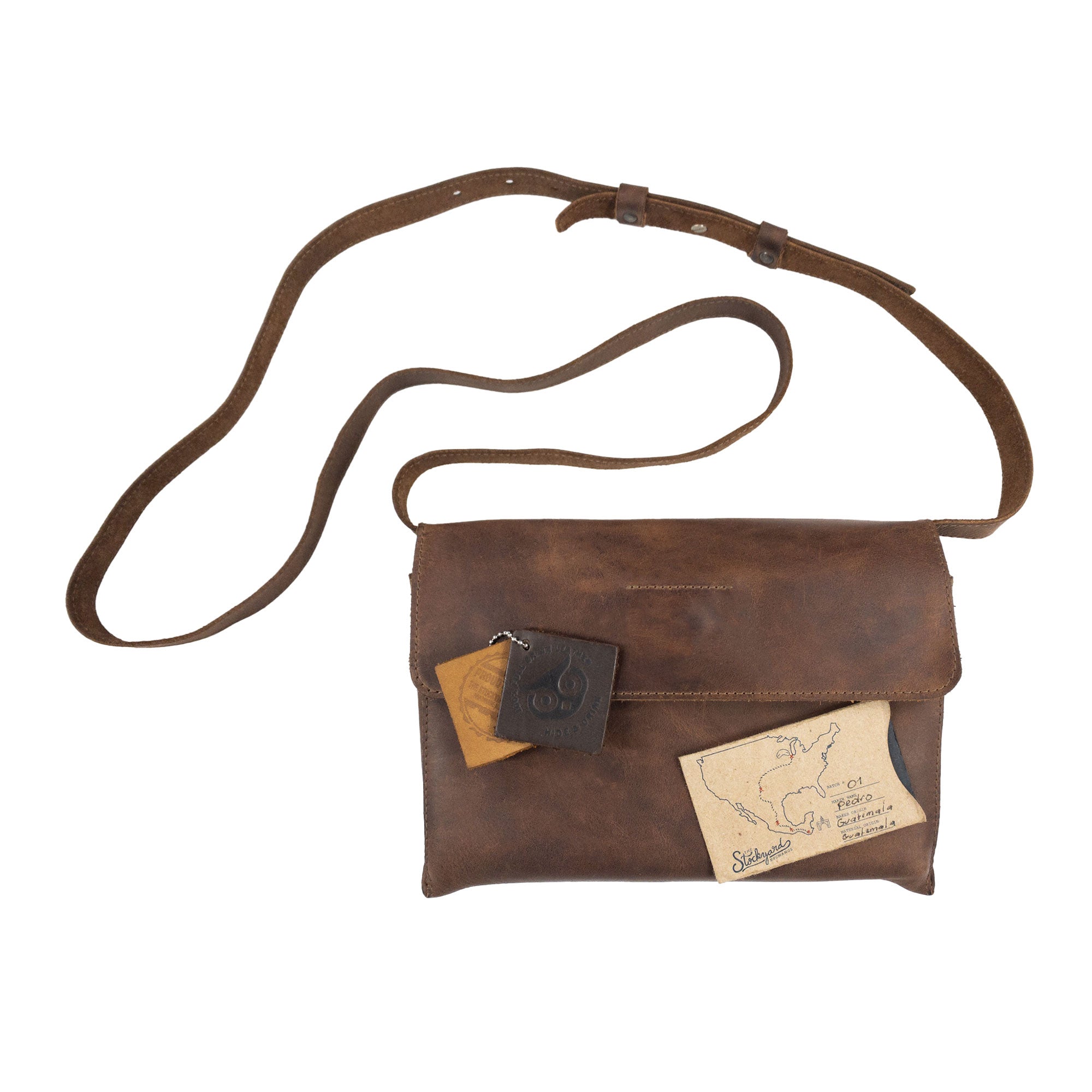 The Minimalist Shoulder Bag by Hide & Drink is styled with a tag and small accessory, showcasing its versatility as a fashionable travel companion. This rectangular messenger bag, made from full grain leather, exemplifies a Mini Classic Shoulder Bag. Crafted through a small batch production process, this artisan piece reflects the meticulous work of hand cut and hand sewn techniques in La Antigua Guatemala & Oaxaca Mexico.