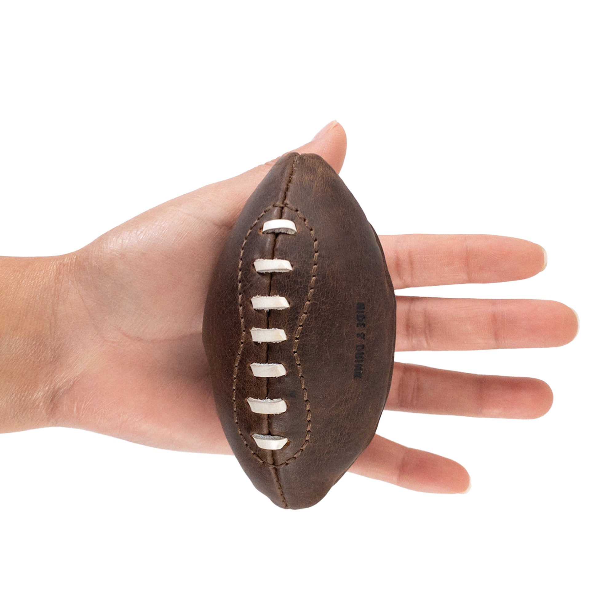 A hand holding a Mini American Football by Hide & Drink, demonstrating its small size and fine craftsmanship. This hand cut ball, made from full grain leather, makes for an excellent keepsake or display item, showcasing the brand's dedication to artisanal production.