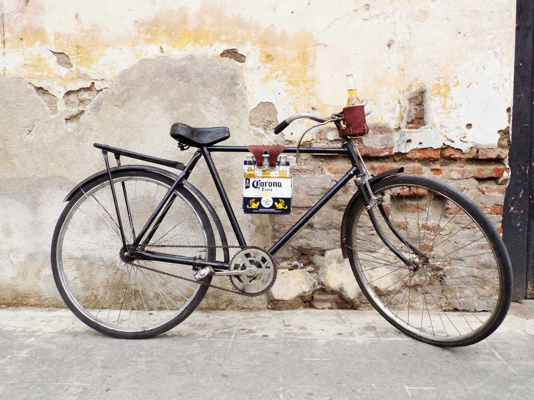 A bicycle adorned with the Hide & Drink Six-Pack Cinch, showcasing a six-pack of Corona beer. This Full Grain Leather Bicycle Beer Carrier emphasizes artisan quality, combining hand cut and hand sewn artistry, handmade with care in La Antigua Guatemala & Oaxaca Mexico.