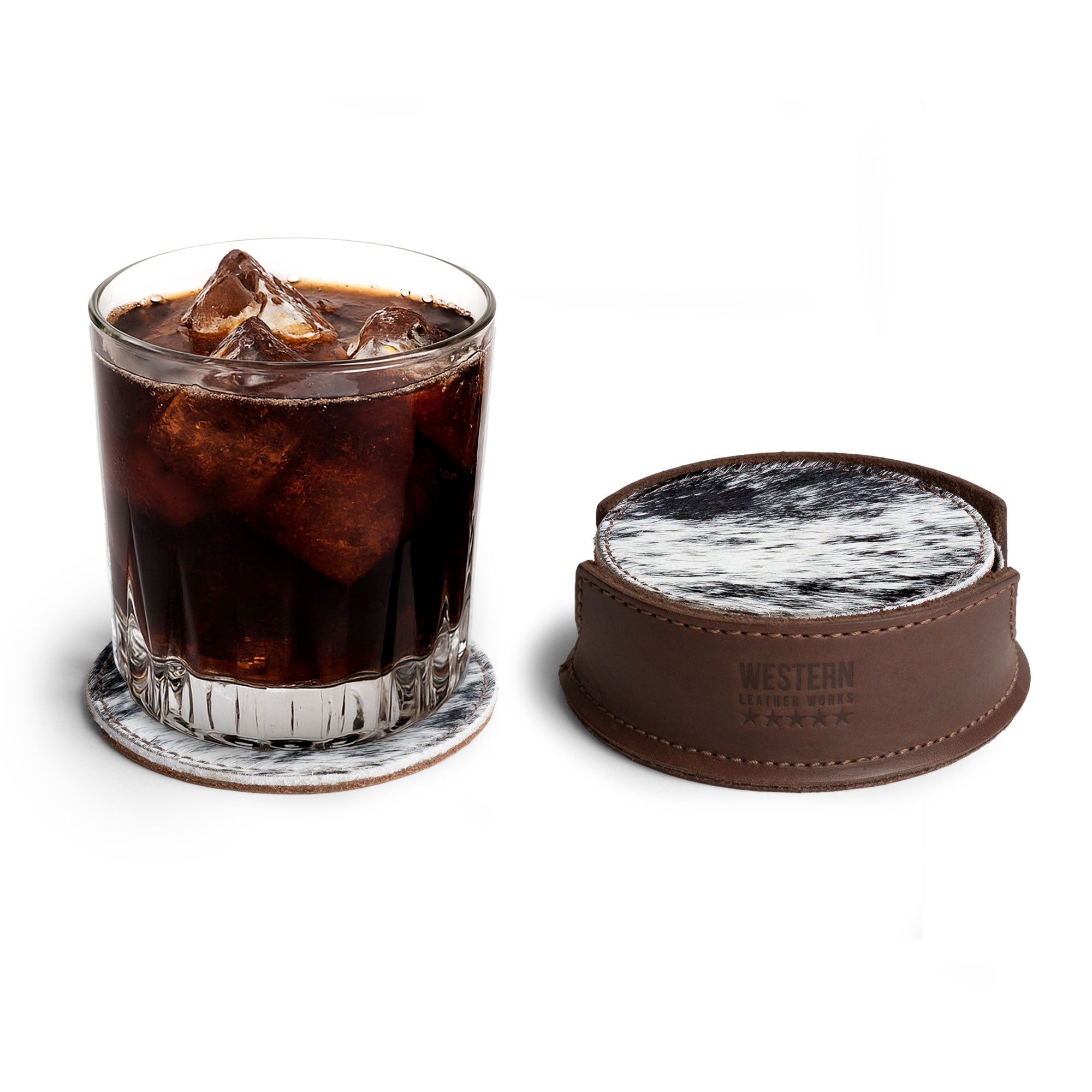 A glass filled with dark iced beverage placed on a handmade hair-on-hide coaster from Western Leather Works, part of the Set of 6 Circular Coasters for Drinks, offering Glass & Cups Stain Protection. This artisan product showcases Full Grain Leather, crafted in small batch production, highlighting the beauty of hand cut and hand sewn techniques, with fire branded logo representing its heritage from La Antigua Guatemala & Oaxaca Mexico.