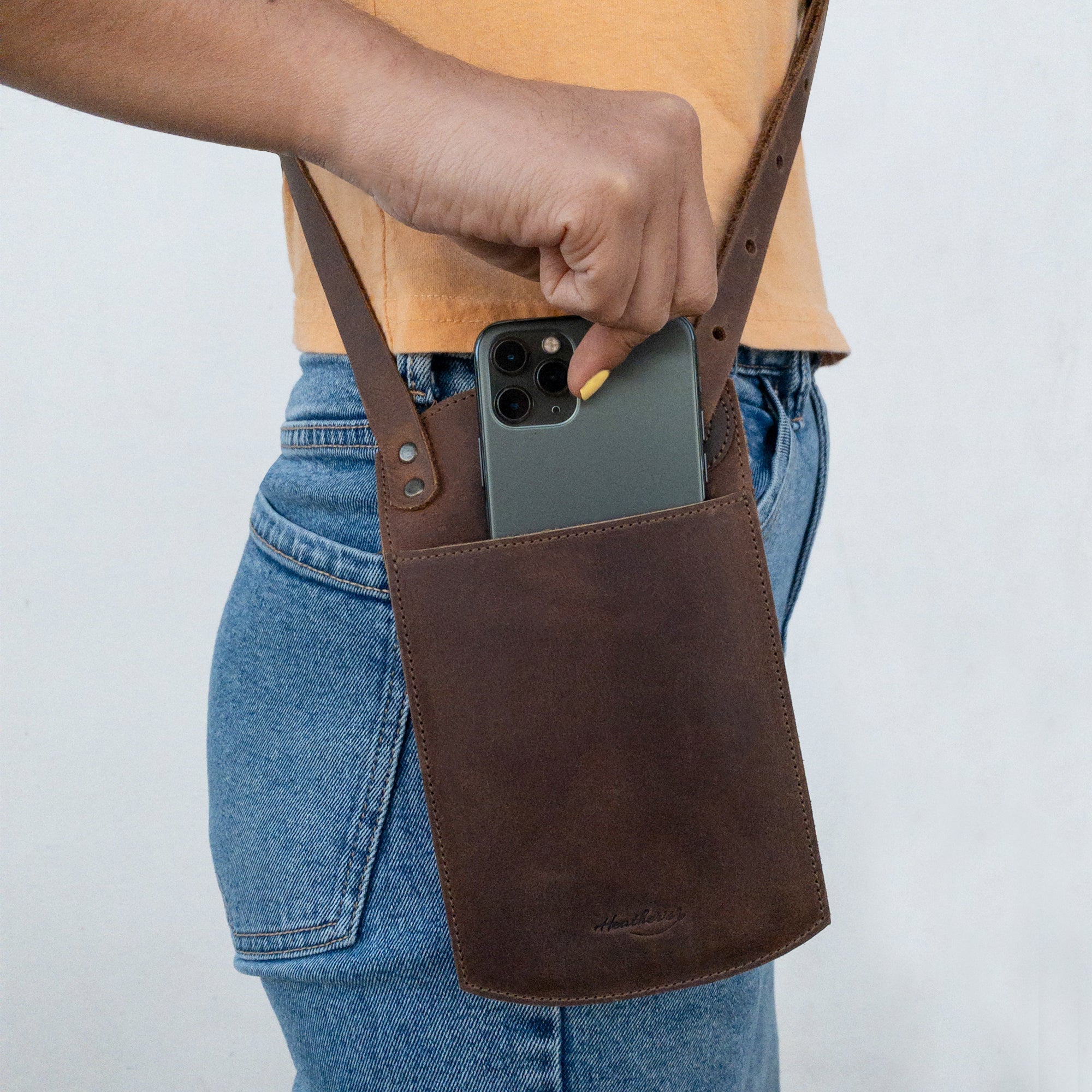 Lifestyle image of a person holding the Phone Purse with Adjustable Strap by Heather's, demonstrating its use as a convenient crossbody bag. This full grain leather bag integrates a horizontal cellphone design with an adjustable neck strap and card slot. Artisan crafted in La Antigua Guatemala, it reflects the brand's commitment to small batch production and handmade quality.
