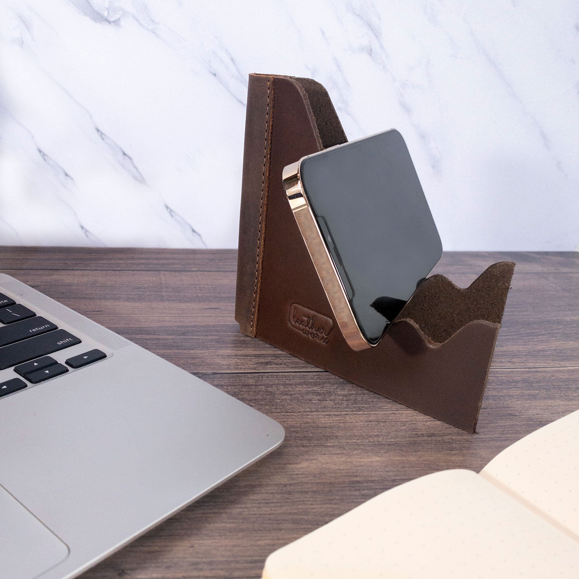 A minimalist Leather Worx Cell Phone Stand for Desk featuring a smartphone resting gently in its foldable design. This rustic Cell Phone Holder serves as a functional office accessory made from full grain leather, representing small batch production and artisan craftsmanship from La Antigua Guatemala & Oaxaca Mexico, with hand cut and hand sewn details.