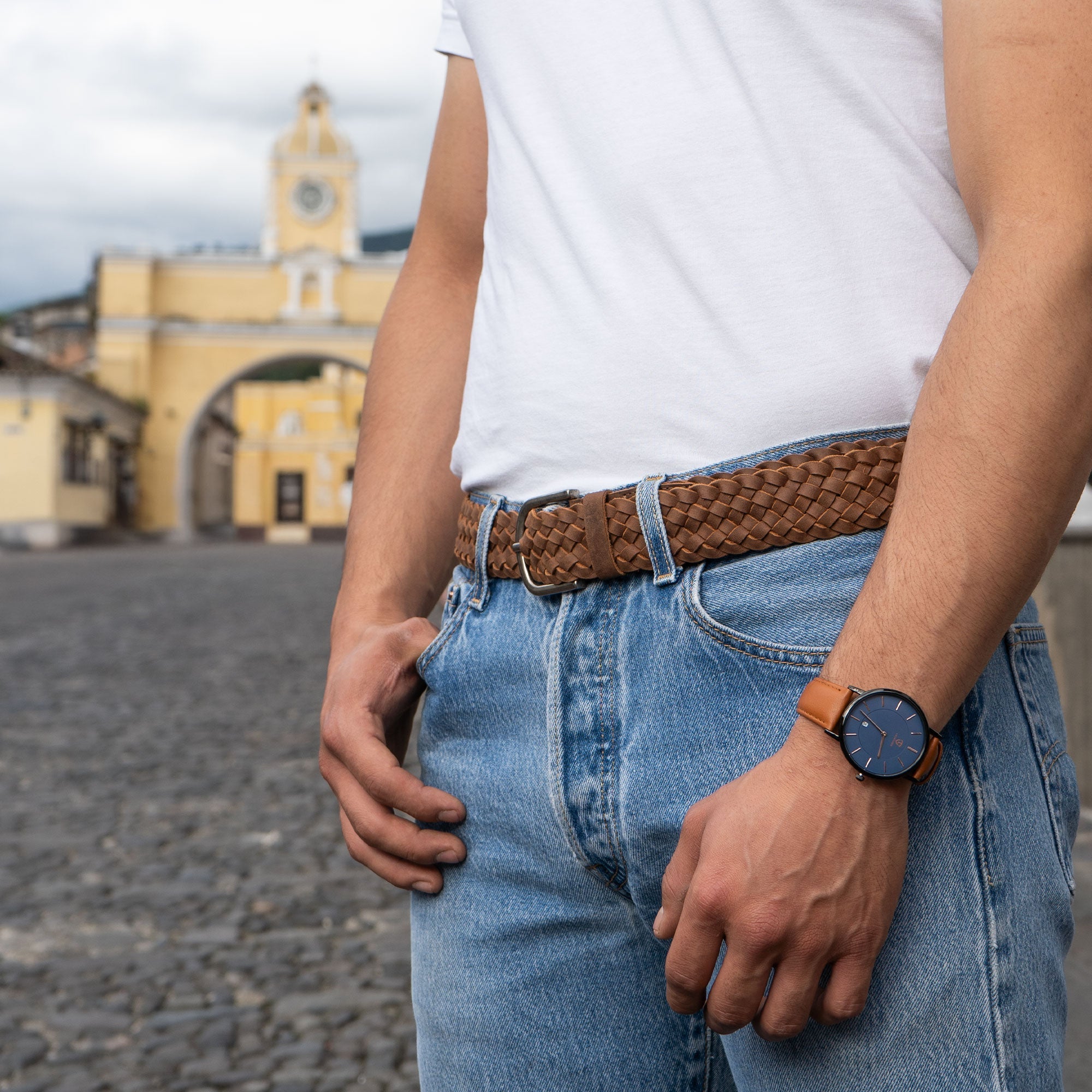 A man wearing the Single-Piece Braided Belt by Hide & Drink, styled with casual denim jeans and a watch against a scenic backdrop. This braided men's belt made of full grain leather is an artisan accessory, reflecting small batch production and the handmade craftsmanship from La Antigua Guatemala & Oaxaca Mexico. It's the perfect combination of style and durability.