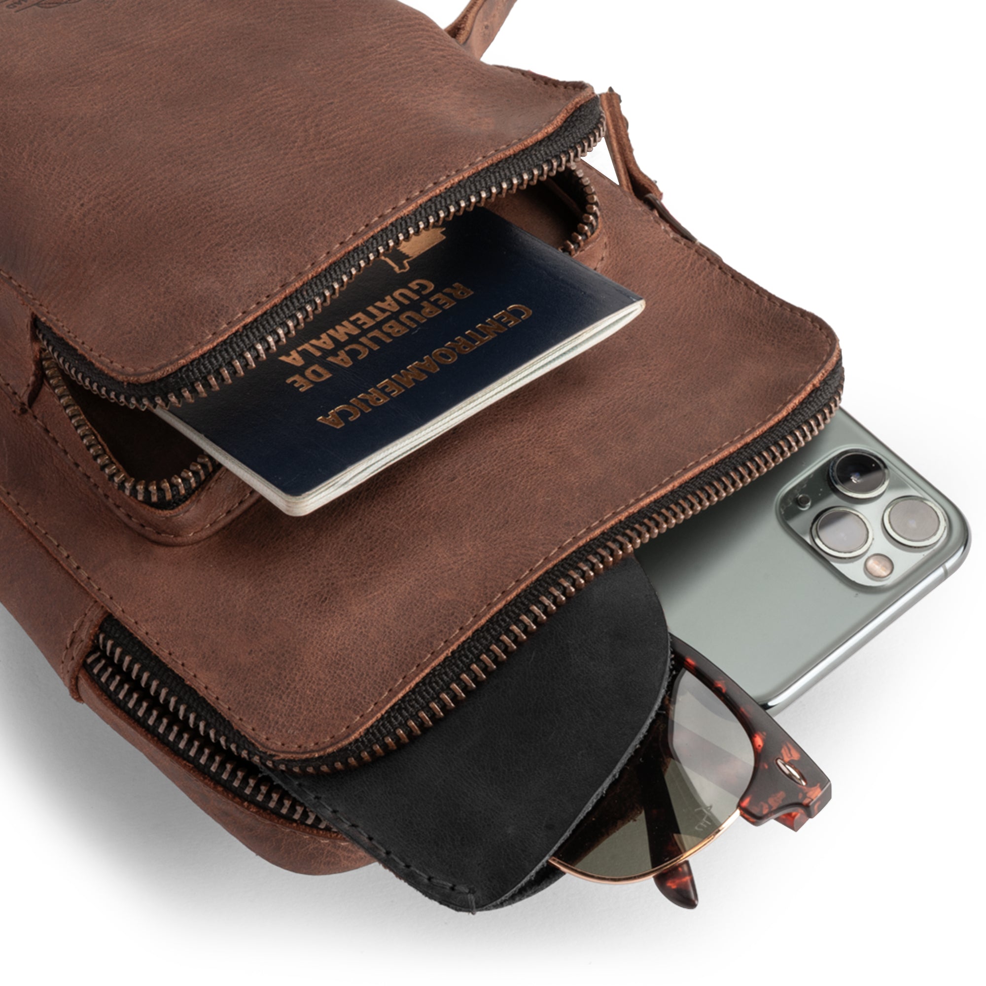 Traveler Sling Bag by Leather Nomad, filled with travel essentials like a passport and phone. This small, trendy bag is made from full grain leather, ideal for men and women on the go. Handmade with care in La Antigua Guatemala, it showcases artisan skills and a stylish design that blends practicality and fashion.