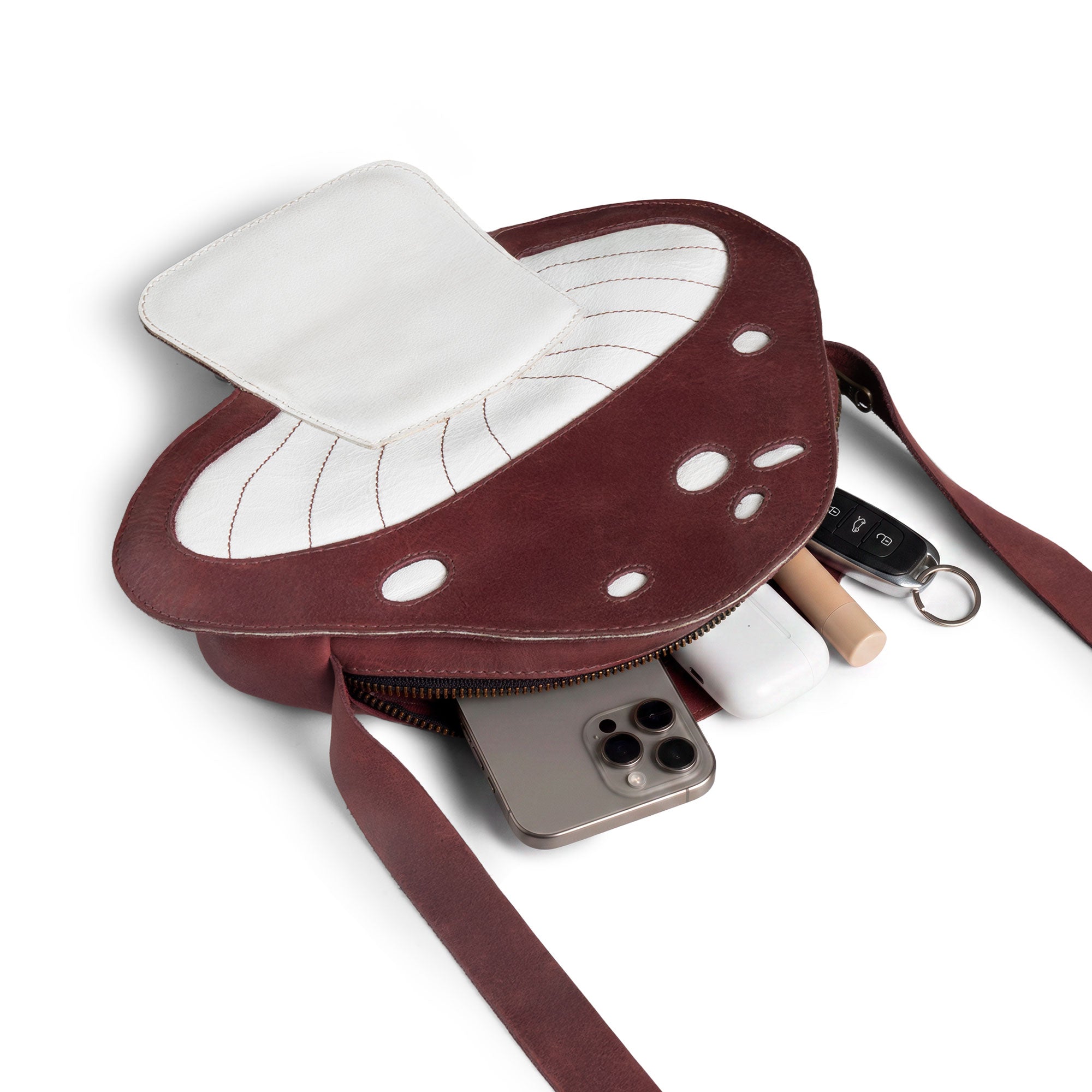 The Mushroom-Shaped Leather Shoulder Bag by Tudi Frudi is shown here filled with everyday items, demonstrating its practicality as a crossbody bag. This playful bag, made from Full Grain Leather, serves well as a travel or messenger bag, reflecting the quality of small batch production with artisan craftsmanship from La Antigua Guatemala.