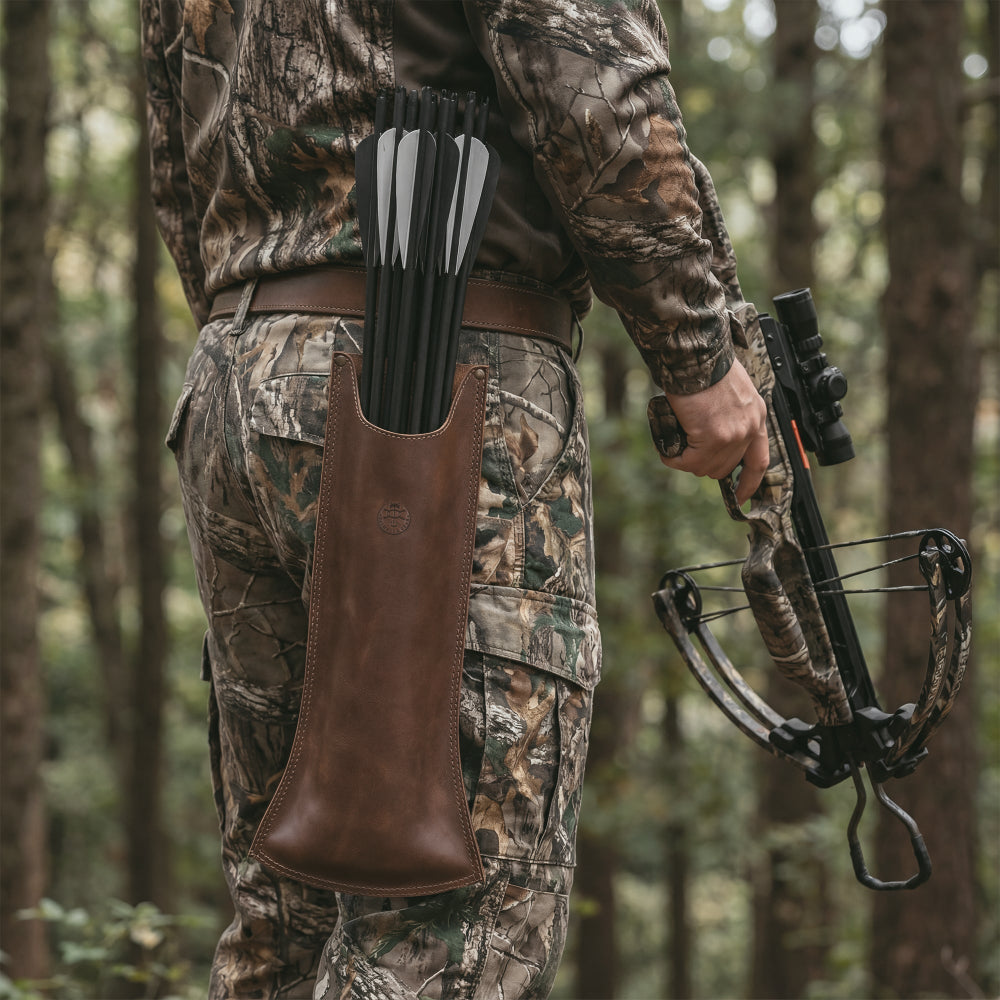 In an outdoor setting, a hunter wears Valhalla Gear's The Fletcher, Hip Bolt Quiver, made from premium full grain leather. This archery accessory is a perfect combination of function and style, serving as a crossbows quiver for bolts and an arrows holder during hunting trips. With its artisan handcrafted touch, this quiver offers a unique mini arrow case solution, ideal for passionate archers.