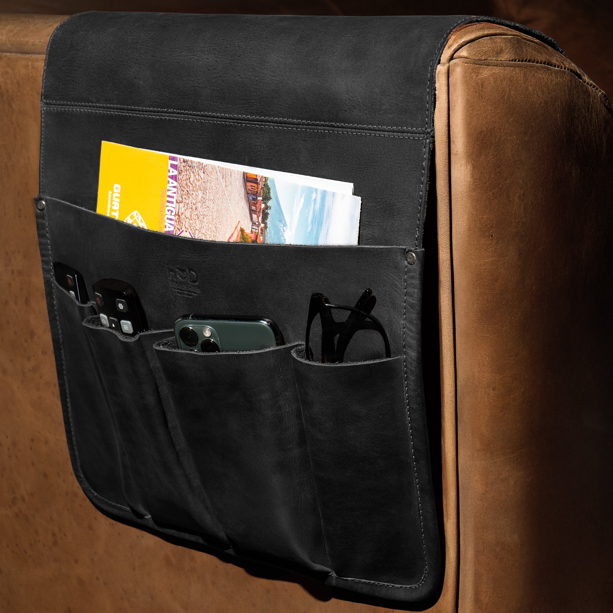 Hide & Drink's Remote Control Holder attached to a couch, elegantly displaying its practical design with pockets for remotes and magazines. A perfect addition to any living room, this armchair caddy is handmade, reflecting the small batch production quality.