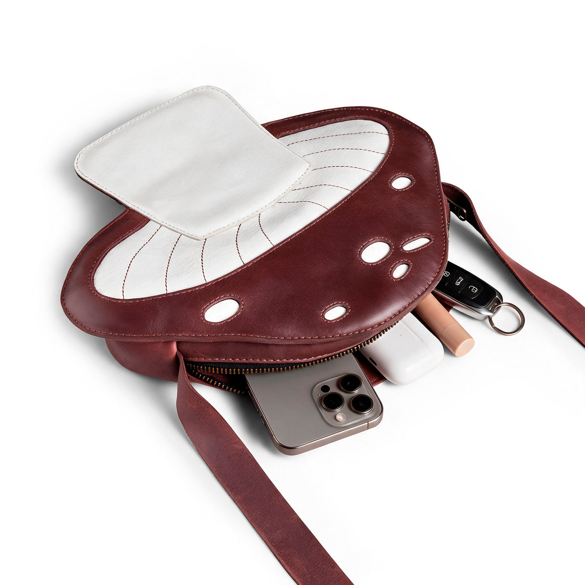 The Mushroom-Shaped Leather Shoulder Bag by Tudi Frudi is shown here filled with everyday items, demonstrating its practicality as a crossbody bag. This playful bag, made from Full Grain Leather, serves well as a travel or messenger bag, reflecting the quality of small batch production with artisan craftsmanship from La Antigua Guatemala.
