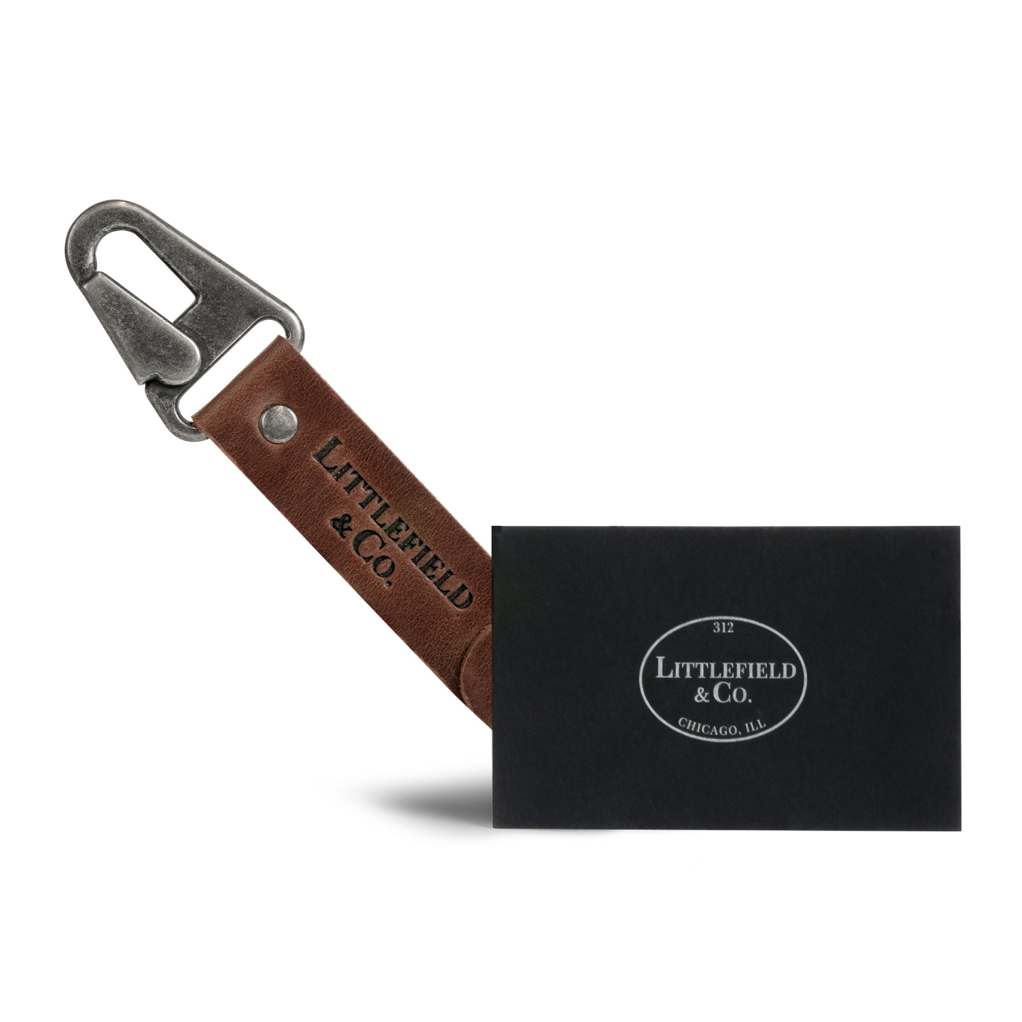 Vintage Adventure Keychain from Littlefield Chicago displayed alongside a business card, illustrating the keychain with clasp and strap with ring design. This rectangular key chain serves as an essential travel accessory for organizing small keys. Crafted from full grain leather, it reflects the artisanal approach of stockyardX with small batch production methods emphasizing hand cut and hand sewn quality, featuring a unique fire branded logo, handmade in La Antigua Guatemala & Oaxaca Mexico.