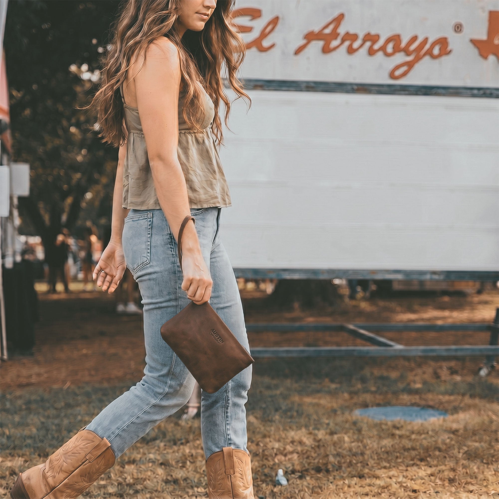 Woman carrying the Austin Genuine The Seaholm Makeup Bag by its leather wrist strap while walking outdoors; the brown Full Grain Leather pouch is part of StockyardX Small Batch Production, Hand Cut, Hand Sewn, Fire Branded, Artisan, Handmade in La Antigua Guatemala & Oaxaca Mexico.