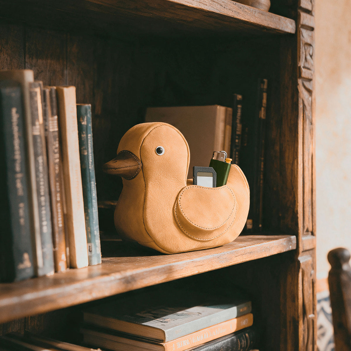 Tudi Frudi Leather Rubber Ducky Pouch displayed on a bookshelf, highlighting its Leather Rubber Ducky Pencil Pouch design, Small Pen Organizer, and Zippered Holder Wallet for Coins, Change Purse, Money Pouches for Men and Women, Saving Box, and Cable Holder functionality; made from Full Grain Leather through StockyardX, Small Batch Production, Hand Cut, Hand Sewn, Fire Branded, Artisan craftsmanship, Handmade in La Antigua Guatemala & Oaxaca Mexico.