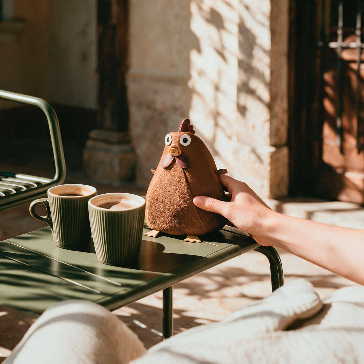 Tudi Frudi Leather Chubby Chicken Pouch displayed on a café table beside coffee cups. This Leather Chubby Chicken Pouch Coin Purse is a Full Grain Leather Small Charger Cable Holder, Change Bag, Scissors Case, and Travel Zippered accessory for Purses & Pouches for Cash, featuring StockyardX Small Batch Production, Hand Cut, Hand Sewn, Fire Branded, Artisan craftsmanship, Handmade in La Antigua Guatemala & Oaxaca Mexico.