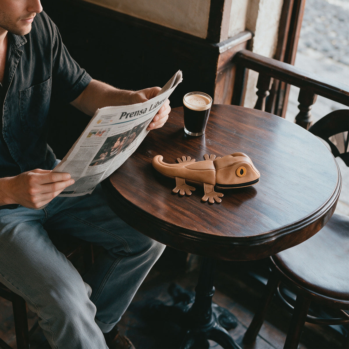Tudi Frudi Gecko Pouch displayed on a cafe table beside a newspaper and coffee, illustrating a portable Gecko Pencil Pouch, Small Pen Organizer, Zippered Holder Wallet for Coins, Change Purse, and Money Pouches for Men and Women; this Full Grain Leather Cable Holder represents StockyardX Small Batch Production, Hand Cut, Hand Sewn, Fire Branded Artisan quality, Handmade in La Antigua Guatemala & Oaxaca Mexico.