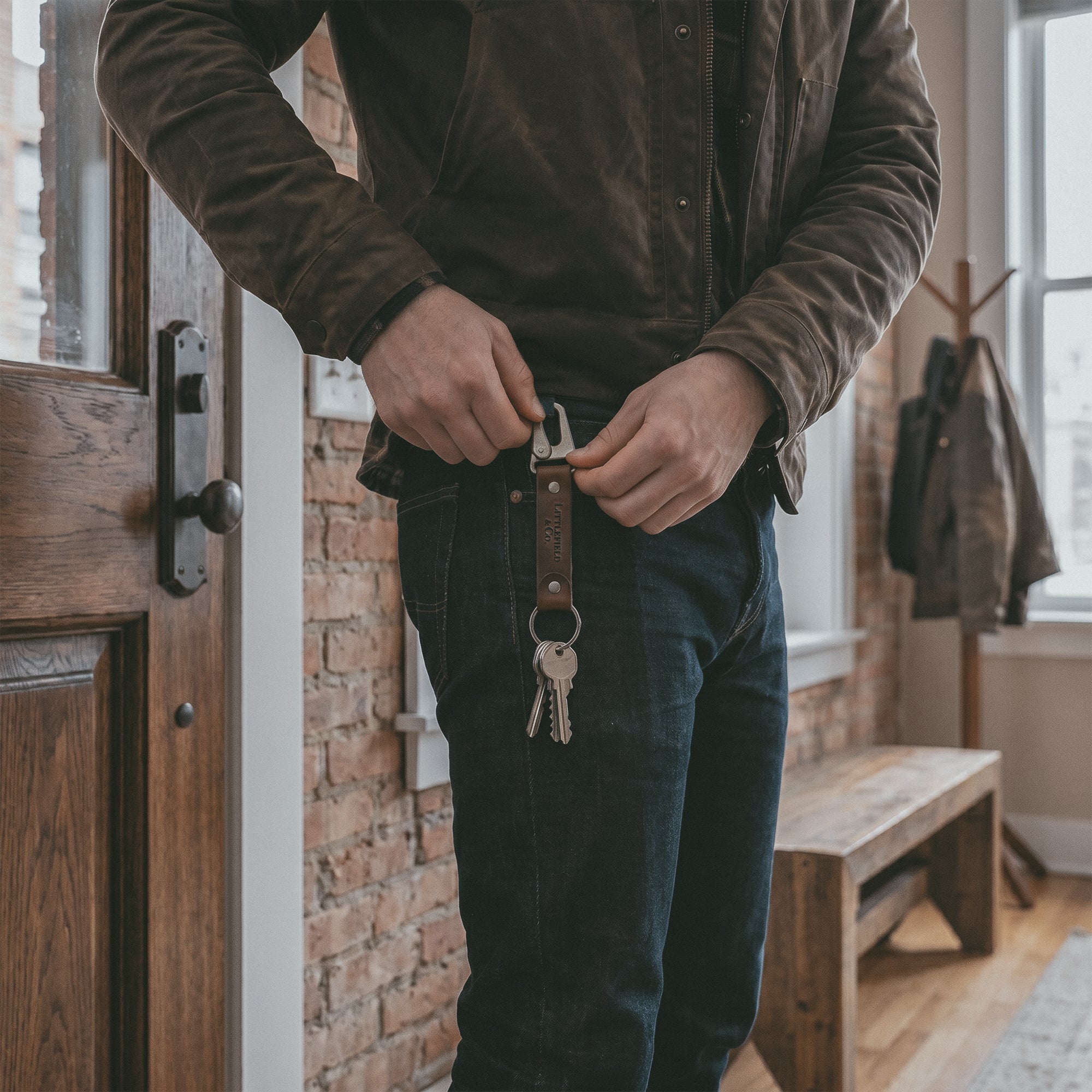 A person using the Vintage Adventure Keychain by Littlefield Chicago with jeans and a jacket, emphasizing the keychain with clasp and strap with ring aesthetic. This rectangular key chain is ideal for men as a travel accessory and small key storage solution. Crafted from full grain leather, it merges style with practicality, embodying stockyardX's dedication to small batch production, hand cut and hand sewn artisanship, and features a fire branded emblem, handmade in La Antigua Guatemala & Oaxaca Mexico.