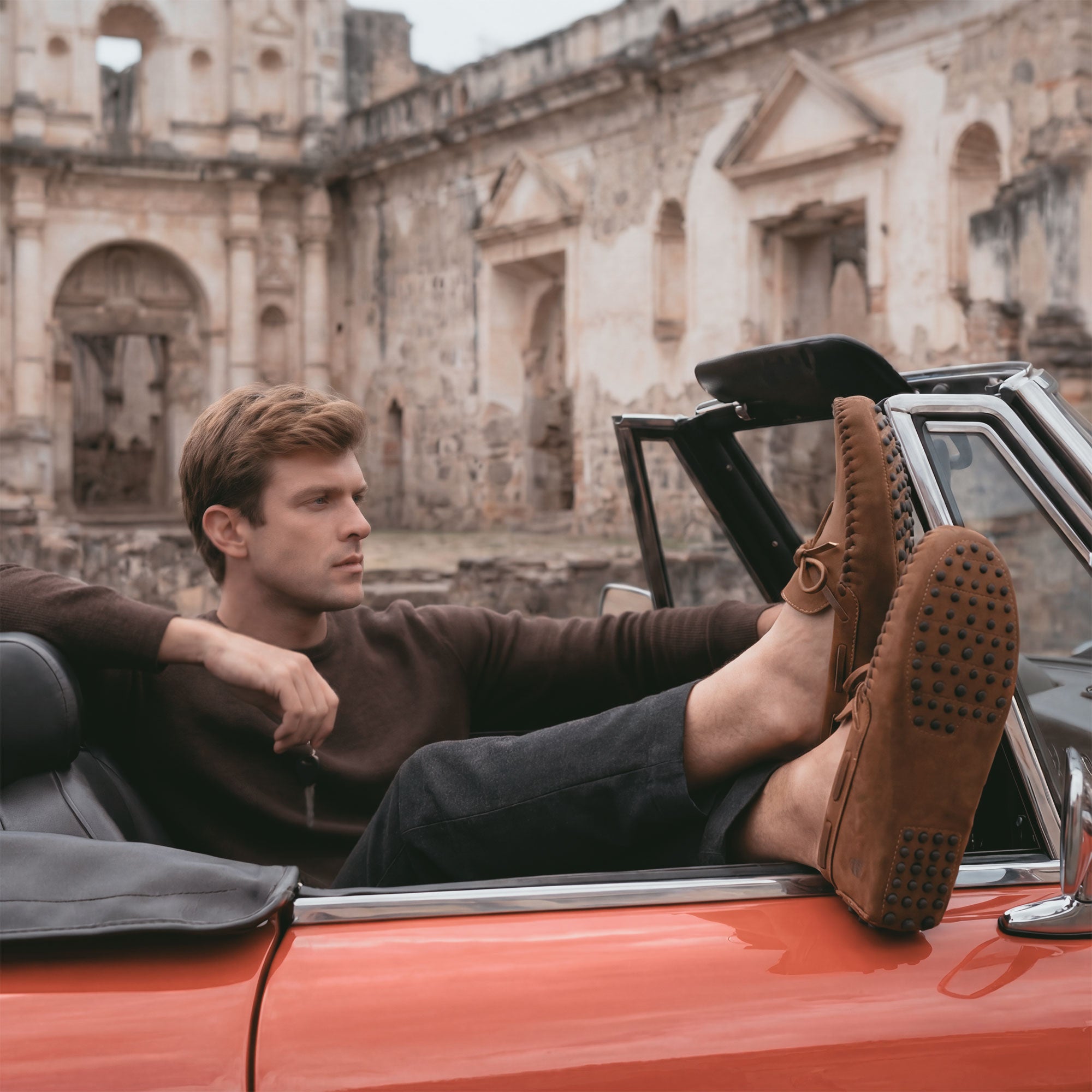 A man enjoying leisure time in The Roadster Leather Driving Moccasins by La Leche, relaxing in a convertible with a view of historic ruins, highlighting the Full Grain Leather and artisan design made in La Antigua Guatemala & Oaxaca Mexico.