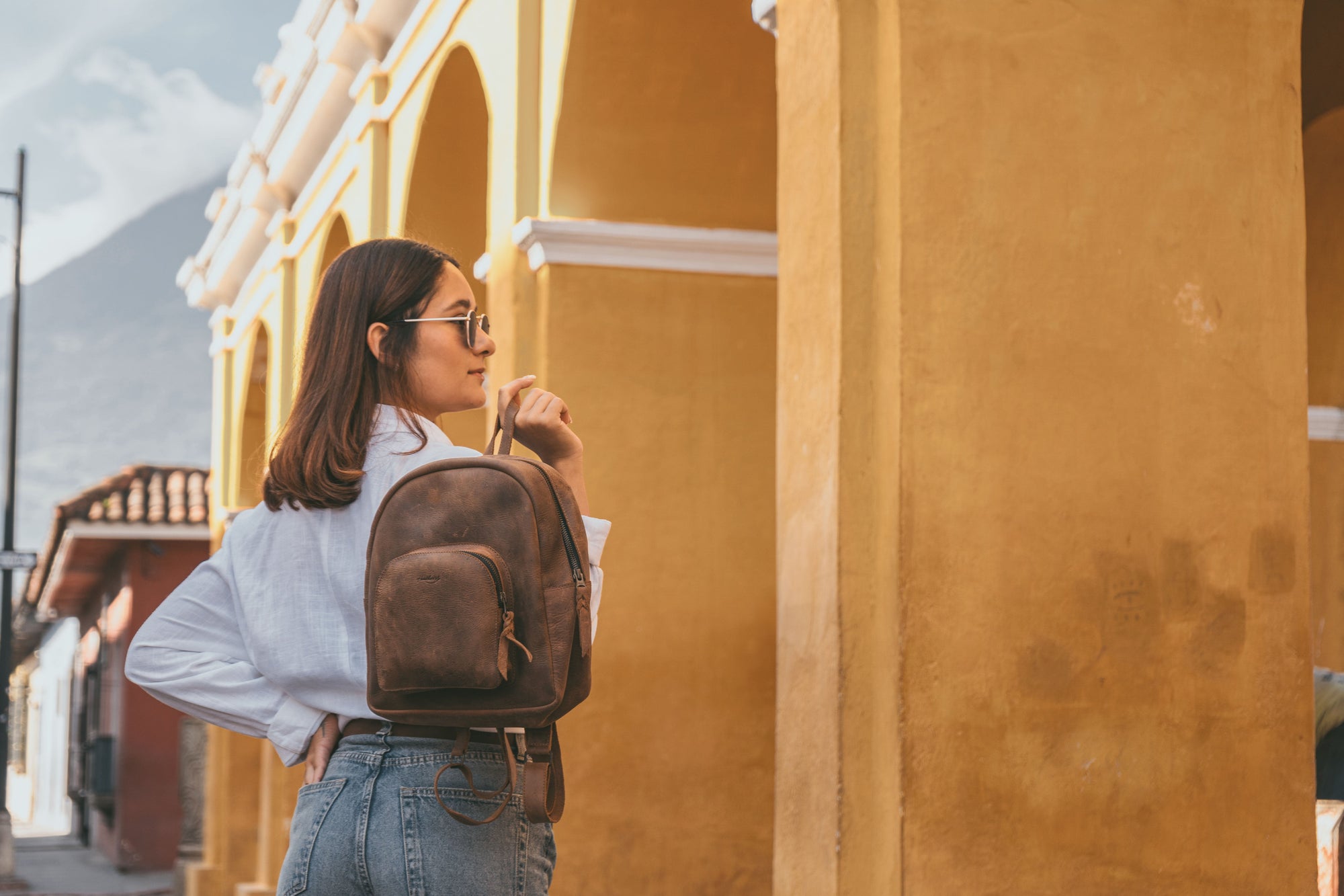 A woman wearing the Heather's Minimalist Backpack against a vibrant yellow backdrop. This stylish full grain leather backpack is perfect for organizing and storing essentials like notebooks and a phone, with adjustable straps for comfort. Handmade in La Antigua Guatemala, this artisan-crafted piece is great for travel and exhibits small batch production quality.