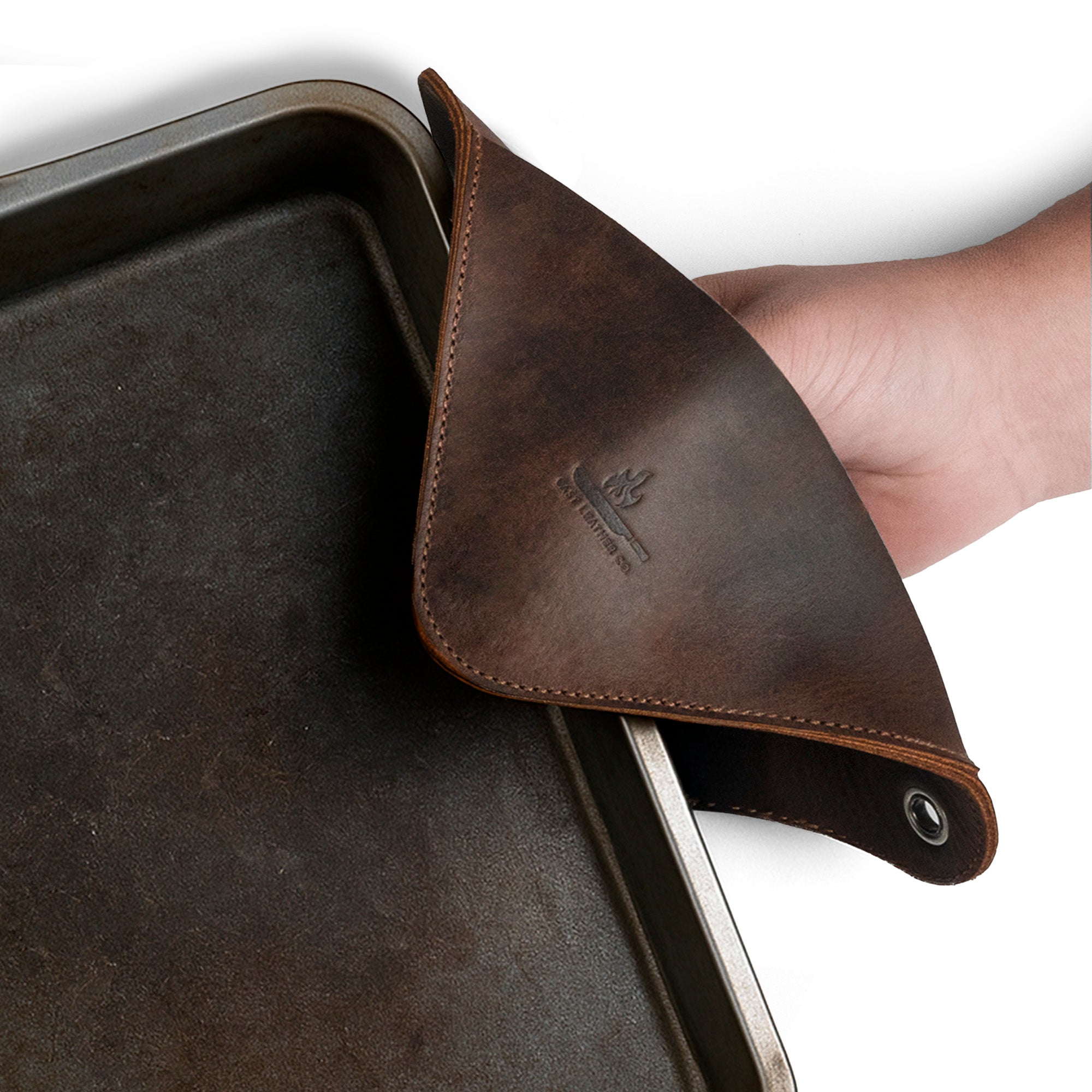 A close-up of a Cast Leather Co. Leather Oven Mitt & Trivet being used to handle a hot baking tray, showcasing its heat-resistant design. This squared hot pot pad (potholder) is made from full grain leather, highlighting its artisan craftsmanship with features like small batch production, hand cut, and hand sewn details, well-suited for bakery supplies. Handmade in La Antigua Guatemala.