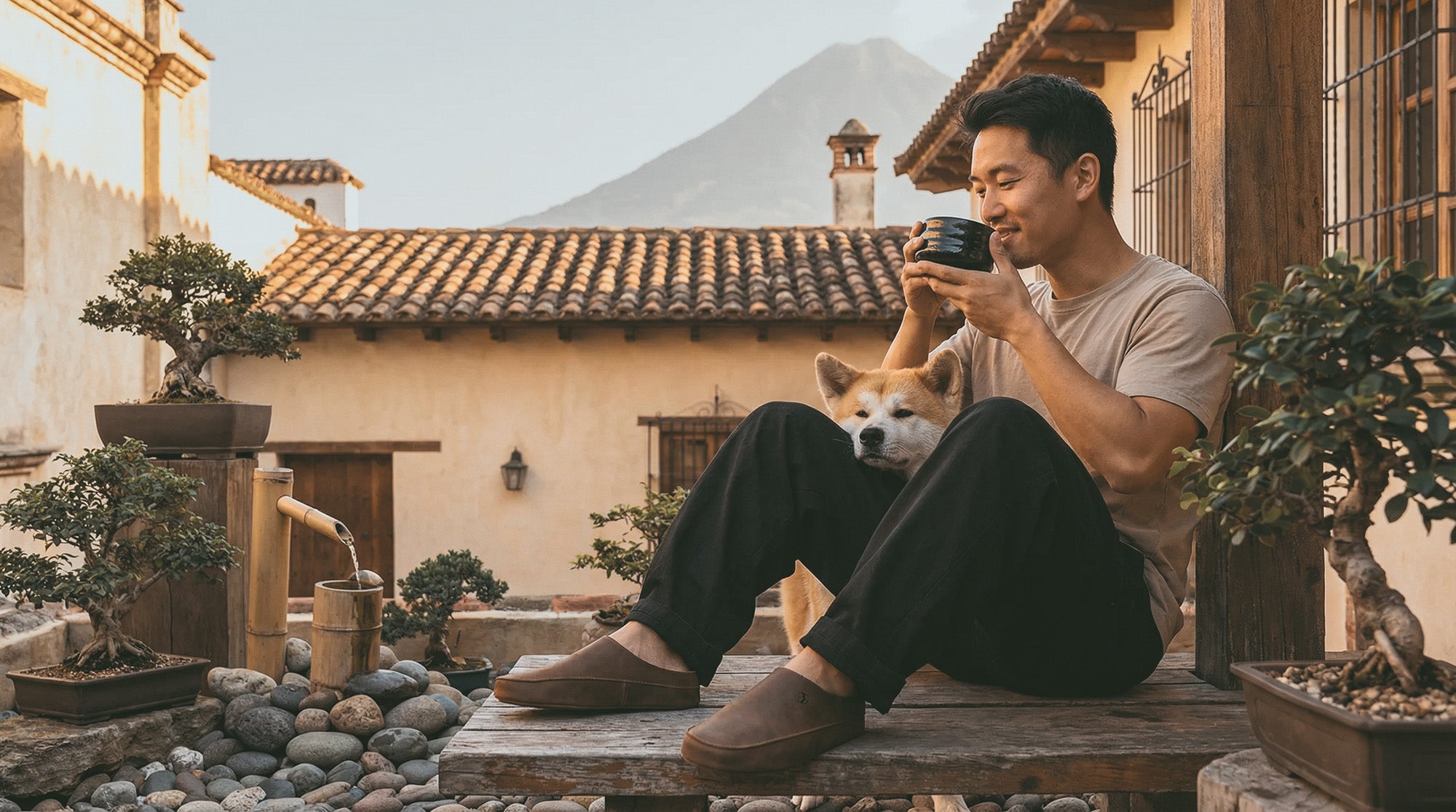 Man sitting on a wooden bench with a dog, holding a coffee cup, in a scenic outdoor setting.