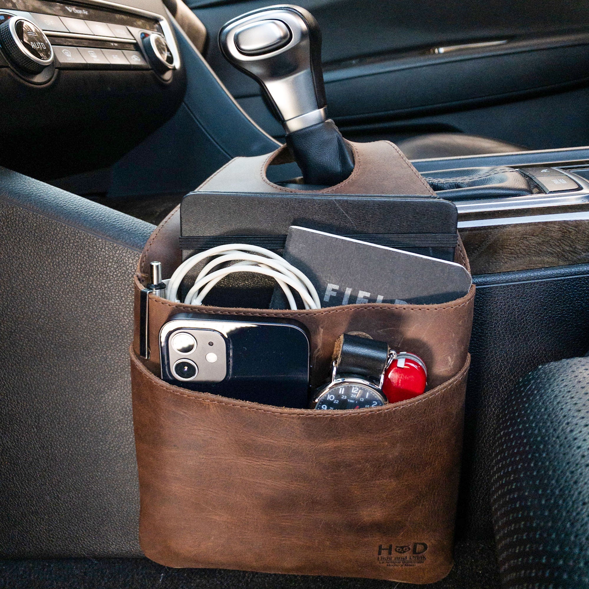Hide & Drink's Seat Side Organizer for Gear Stick in a car setting, showcasing its functionality in Full Grain Leather. The organizer holds chargers, cables, and essentials while protecting outdoor tools like knives. This artisan piece highlights small batch production techniques, being hand cut, hand sewn, and fire branded in La Antigua Guatemala & Oaxaca Mexico.