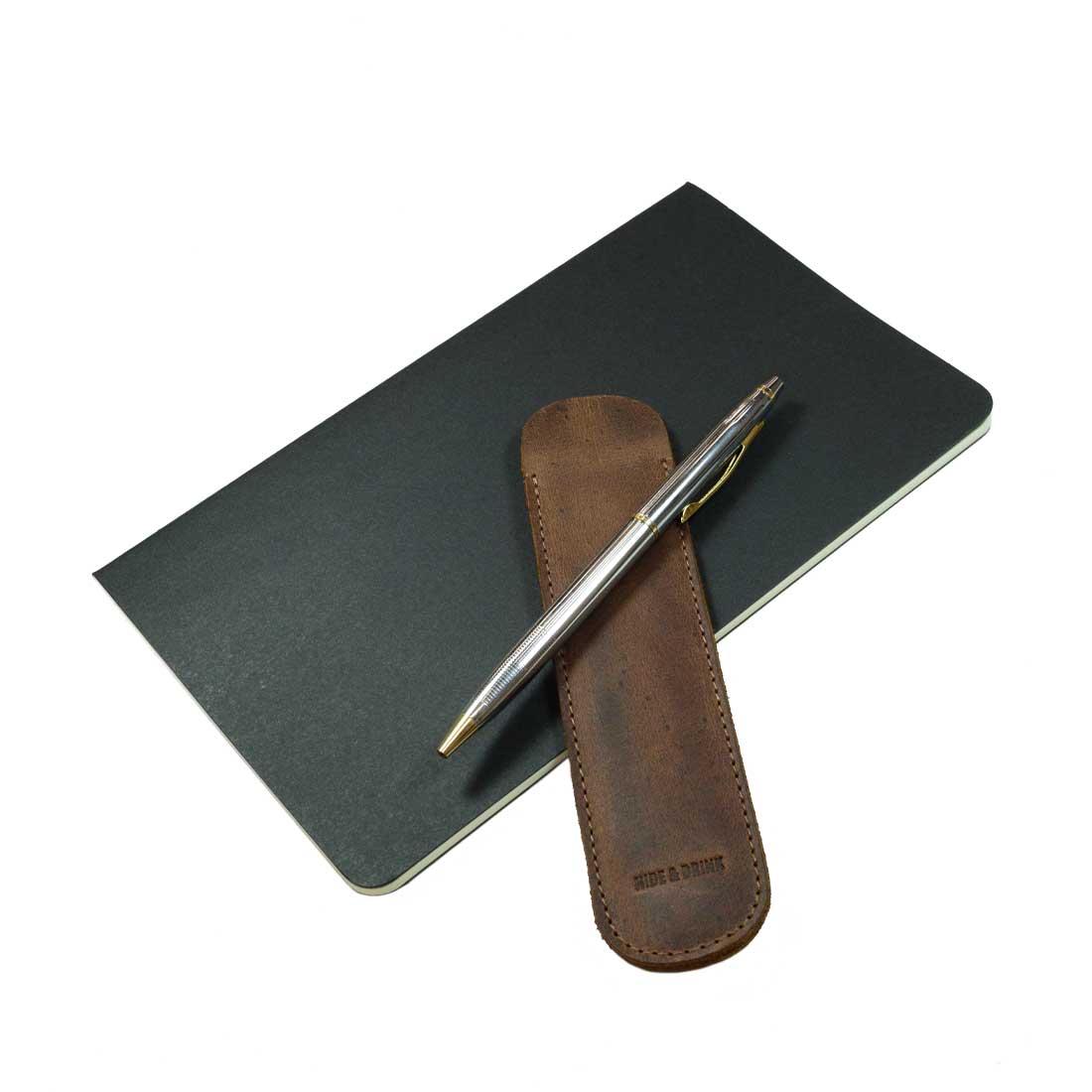 The Vintage Pen Sleeve from Hide & Drink rests alongside a notebook, emphasizing its stylish and functional design as an essential accessory for any bookworm. Made from high-quality Full Grain Leather, each piece is hand cut and hand sewn.