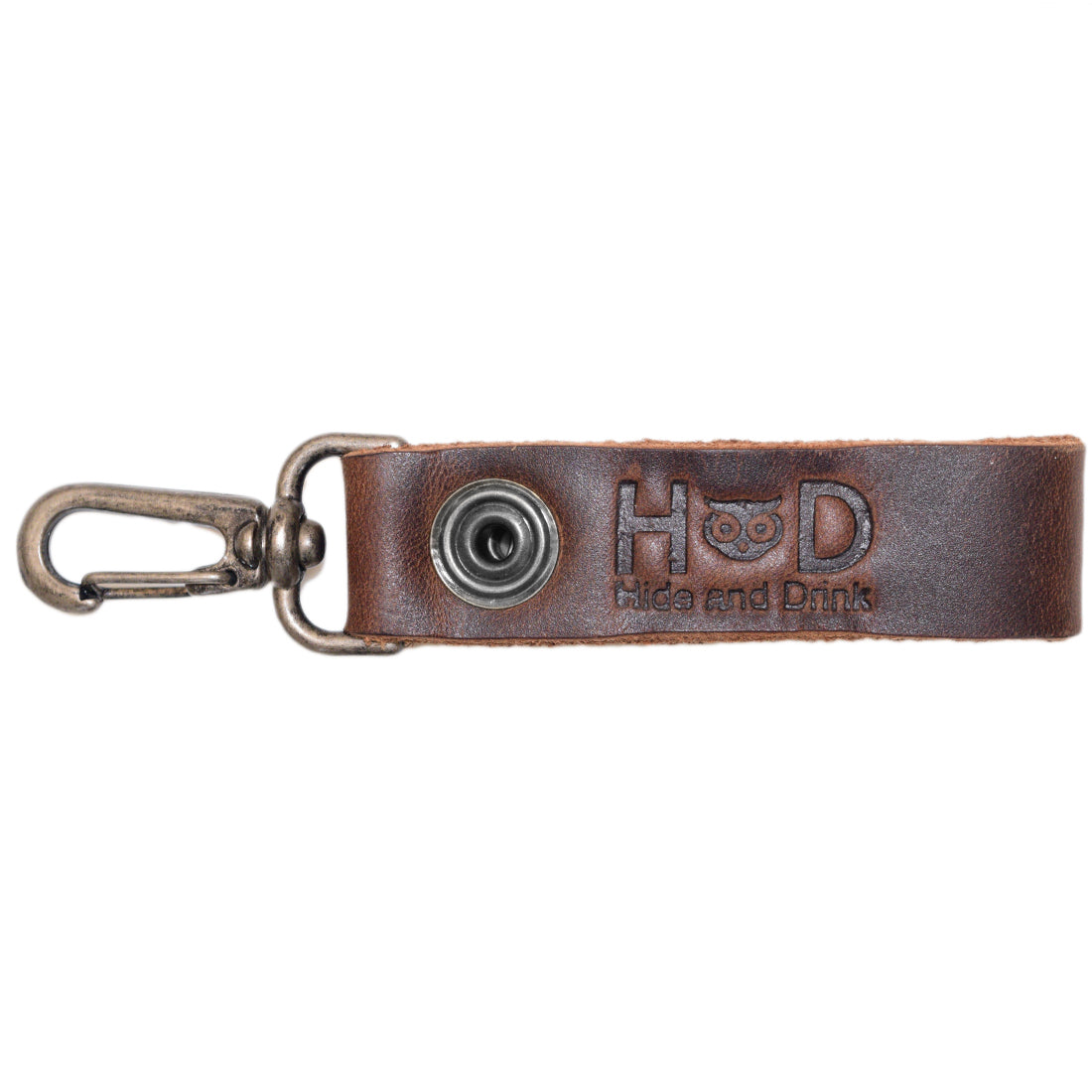 Belt Key Holder by Hide & Drink showcasing the brand logo with a sturdy metal clip, perfect for everyday use. Dimensions Length: 4.25 in. Height: 1 in. Width: 0.50 in. The Full Grain Leather will acquire a distinct patina with usage; conditioning can be done with mineral oil or beeswax.