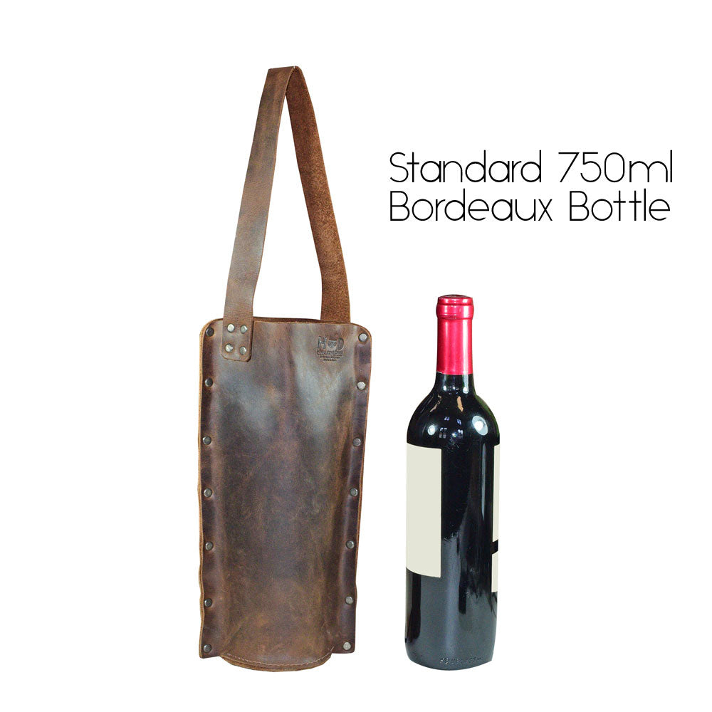 Rustic Wine Carrier For Standard 750ml Bordeaux Bottle by Hide & Drink, pictured next to a standard 750ml Bordeaux bottle. This Single Wine Carrier is made from Full Grain Leather, representing artisan craftsmanship through small batch production, hand cut, hand sewn, fire branded, and handmade in La Antigua Guatemala & Oaxaca Mexico.